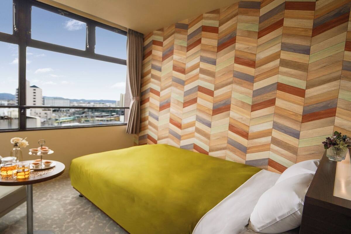 A hotel room with a large window, a bed with a green cover, a small table with a tray of snacks, and a view of a cityscape.