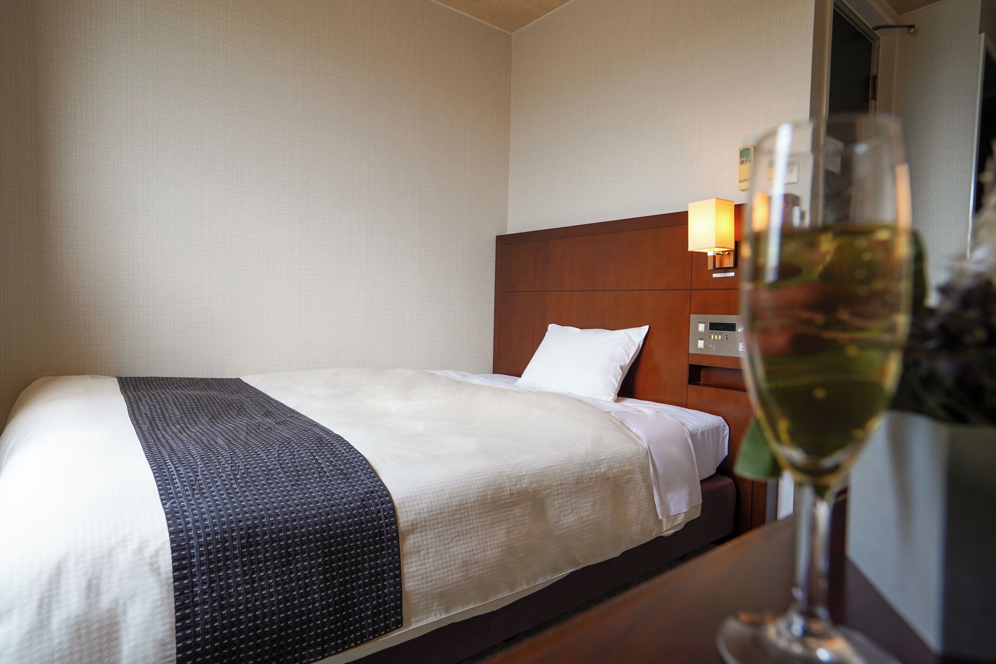 A hotel room with a neatly made bed, a bedside table with a glass of wine, and a lamp on the nightstand.