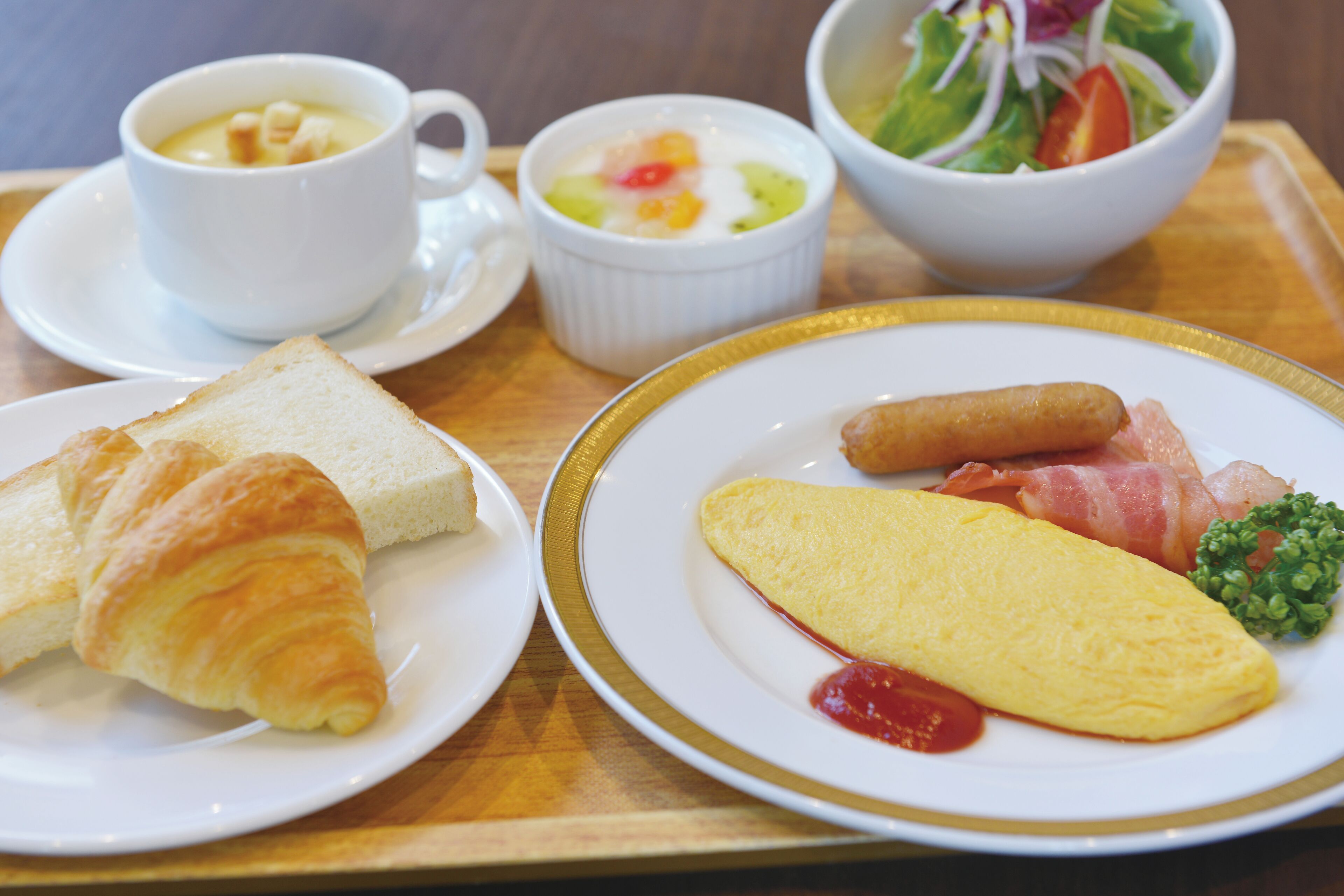 A tray with breakfast items including croissants, an omelette, sausages, and a bowl of salad.