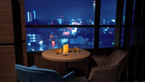 Una habitación con una mesa redonda de madera, dos sillas, una vela y vasos con una bebida. Un ventanal ofrece vistas de un paisaje urbano nocturno.