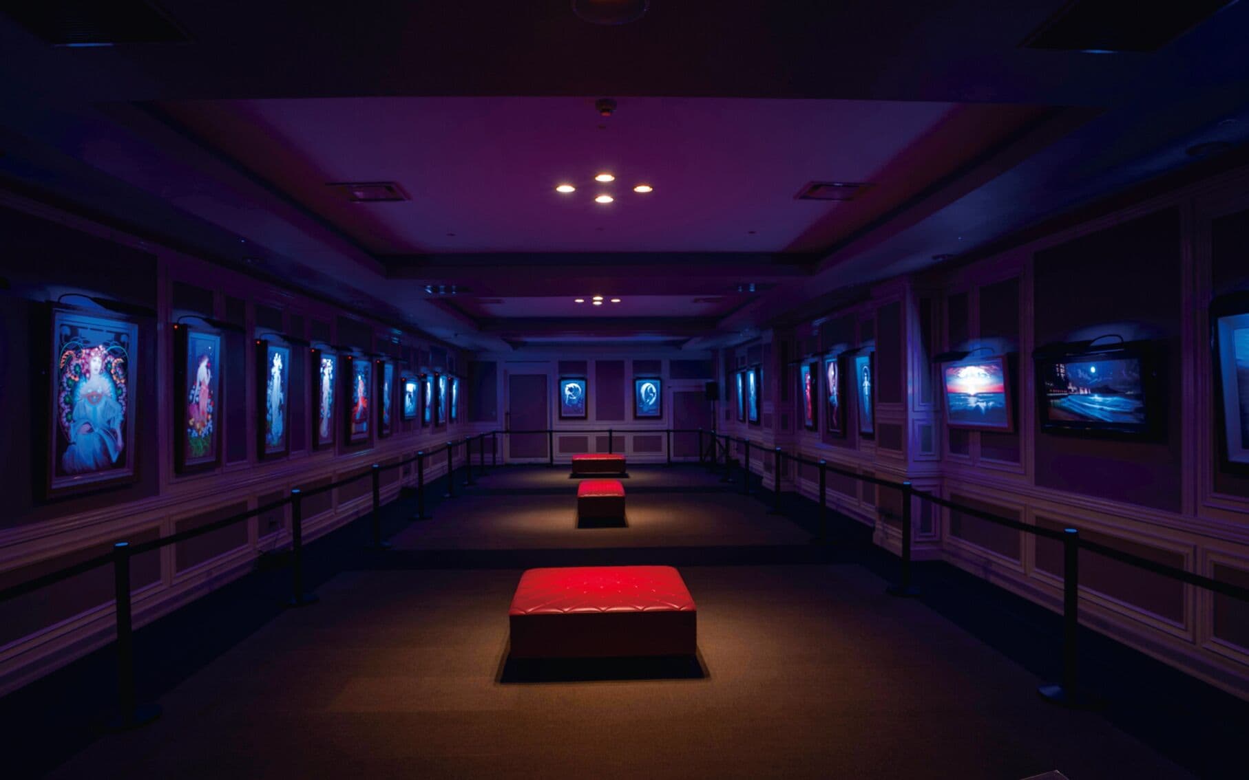 A hallway with framed pictures on the walls, a red bench in the center, and a ceiling with recessed lighting.