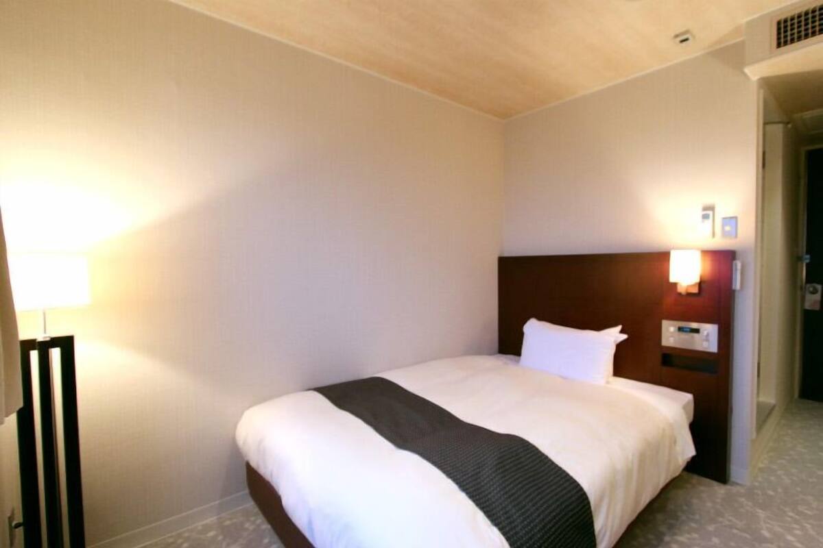 A hotel room with a single bed, a bedside table, a lamp, and a wall-mounted light fixture.