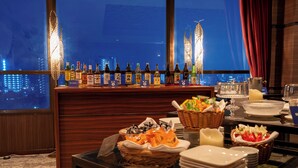 Zona de bar con botellas de licores, bufé con diversos platos y vistas a la ciudad a través de amplias ventanas.