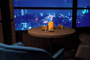 A round table with two glasses of amber liquid, a candle, and a plate, set against a cityscape view through large windows. - Hotel New Gaea Omuta Garden (Omuta)