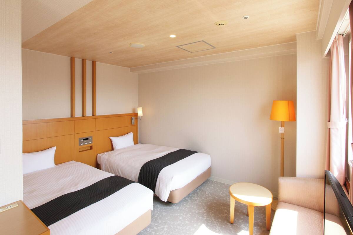A hotel room with two beds, a small table, a lamp, and a window with curtains.