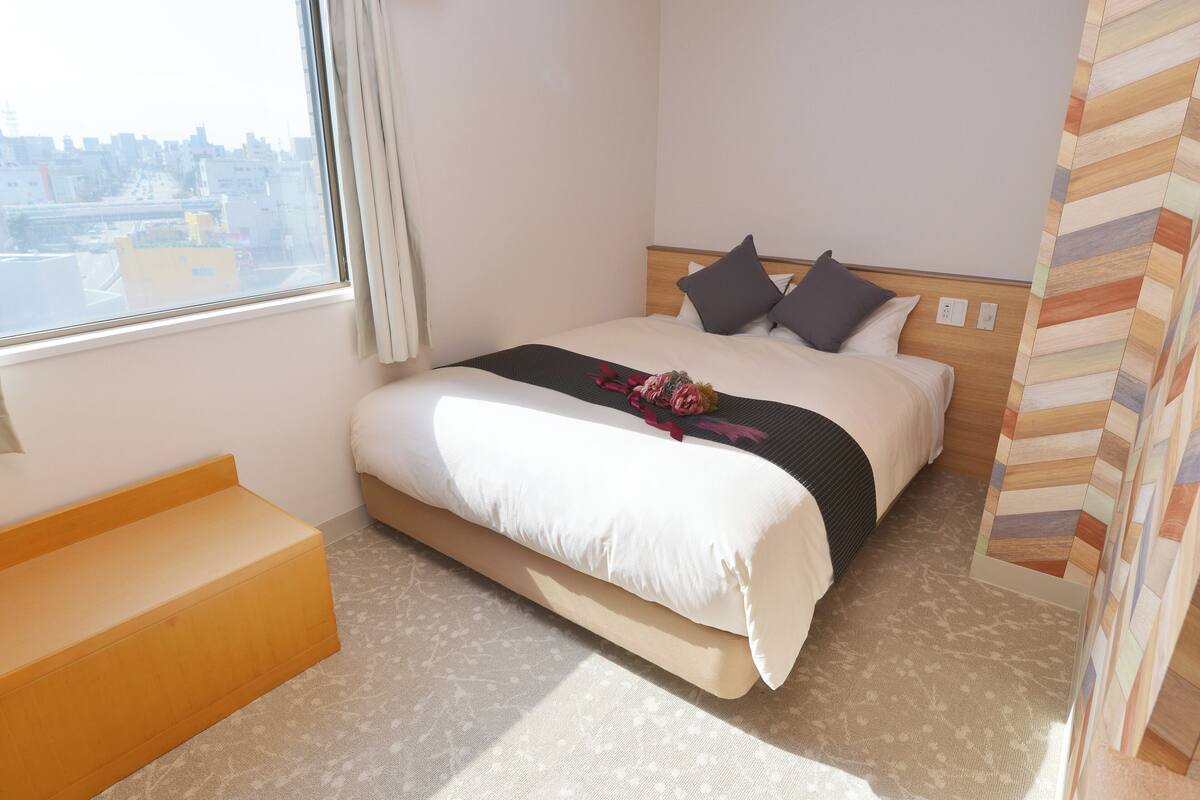 A hotel room with a bed, a bench, a window with a view of the city, and a patterned wall.