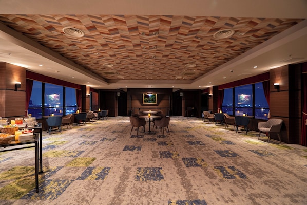 A hotel lobby with a patterned carpet, a central dining area with tables and chairs, and large windows offering a view of the cityscape at night. - Hotel New Gaea Omuta Garden (Omuta)