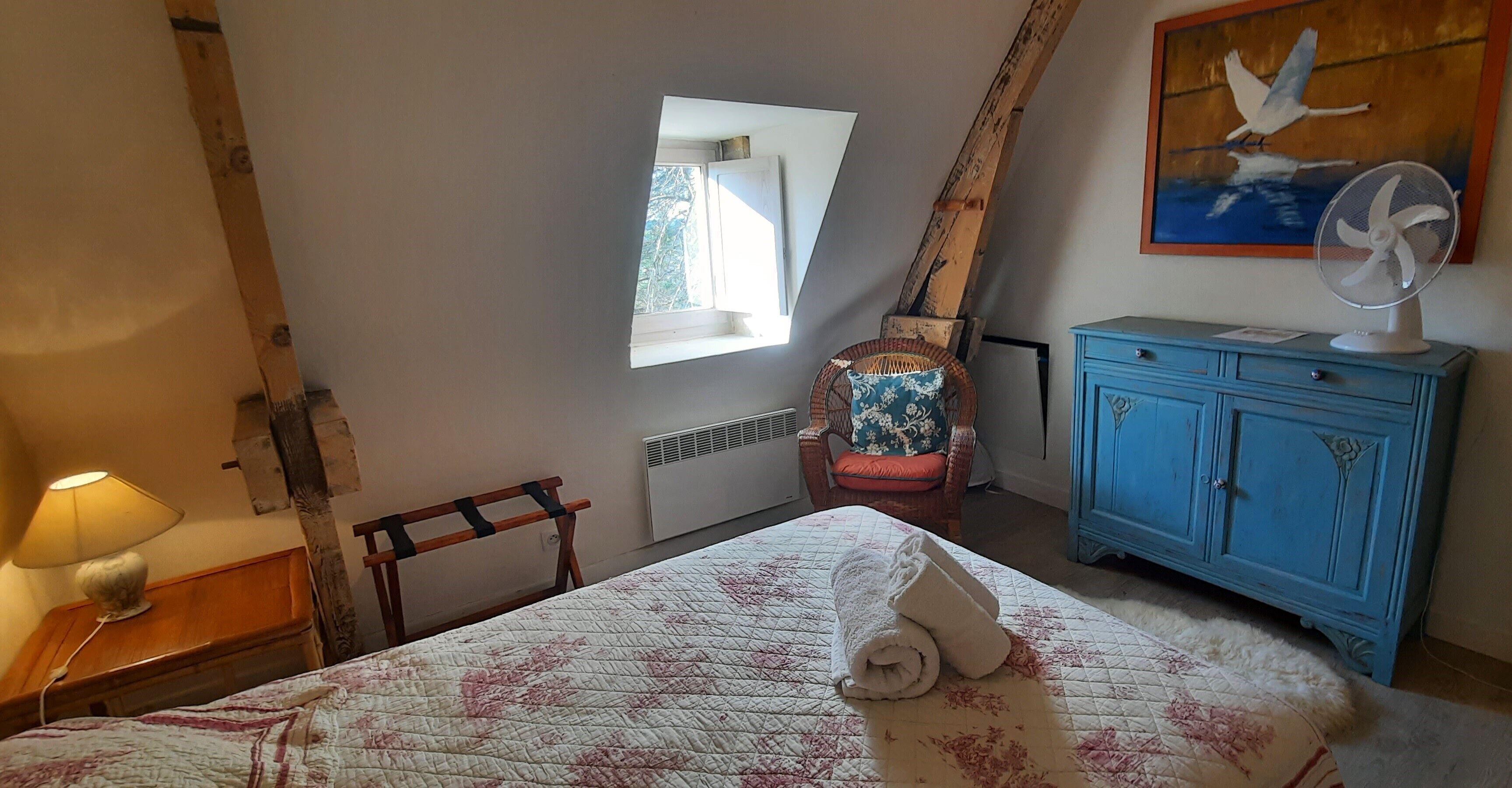 A bedroom with a bed, a window, a chair, a fan, and a blue cabinet.