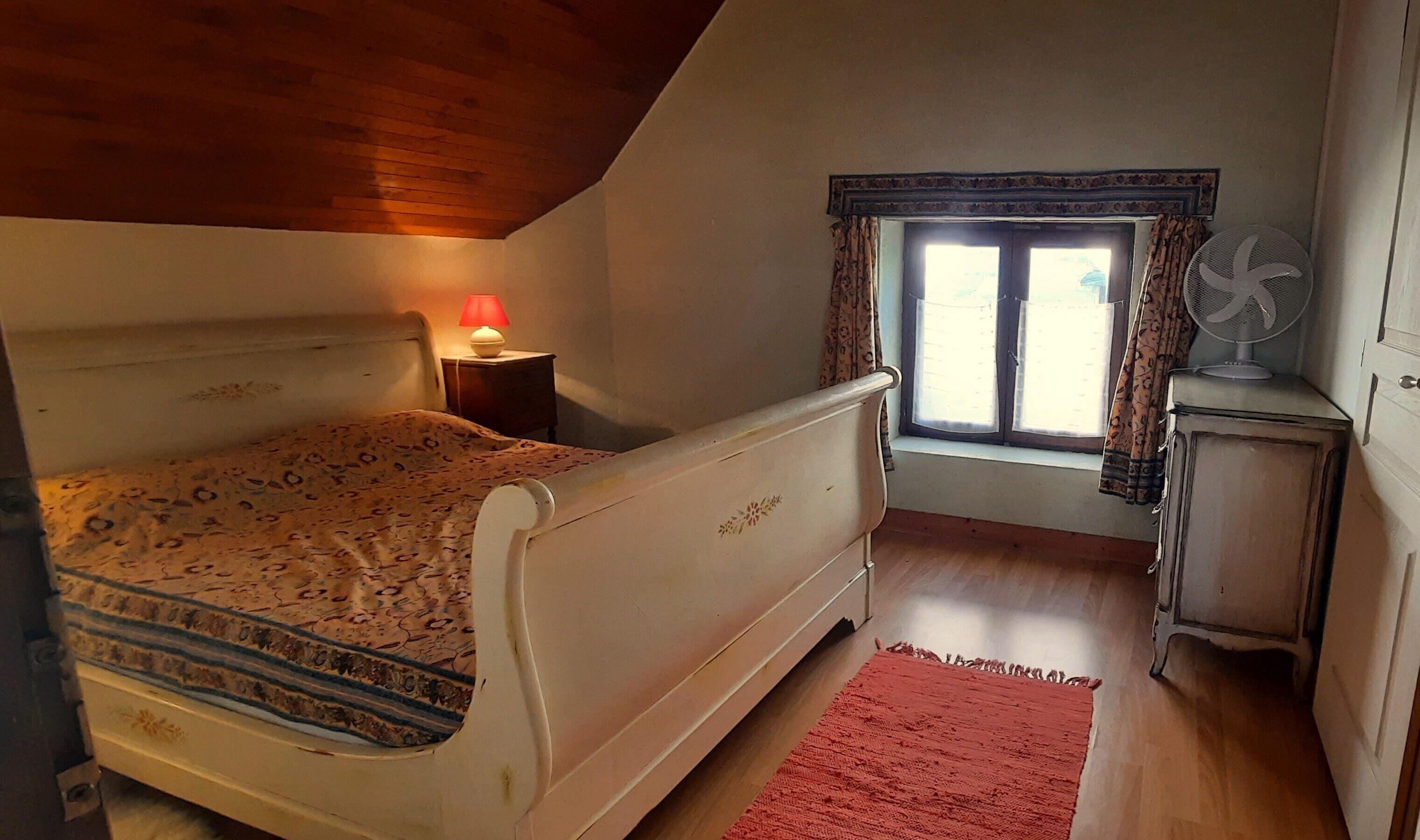 A bedroom with a bed, a nightstand with a lamp, a window with curtains, a radiator, and a small cabinet.
