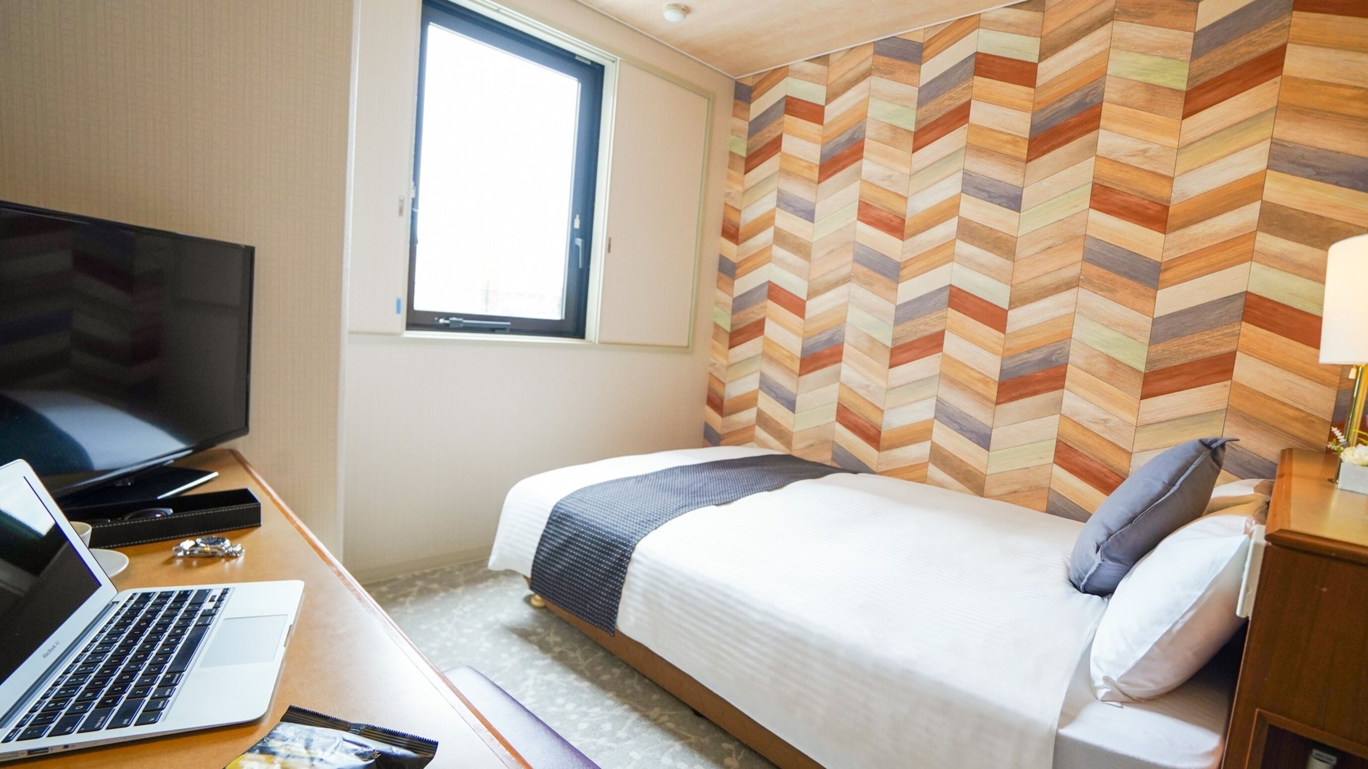 A hotel room with a bed, a desk with a laptop, a TV, and a window.