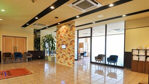 Un hall d’hôtel avec un pilier orné d’un motif en chevrons, un comptoir d’accueil, plusieurs chaises, une plante et un sol carrelé.