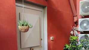 A red-walled entrance with a grey door, a hanging wicker basket with plants, and a potted plant beside the door. - Piazzetta 4 - Holiday Home (Napoli)