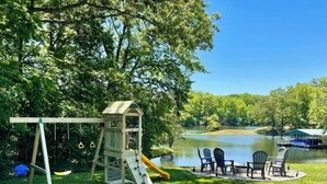 Un parque infantil con tobogán y columpios, una mesa de picnic con sillas, y vista al lago.
