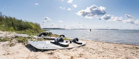 Een zandstrand met een surfplank en een paar koptelefoons in het zand, met uitzicht op een wateroppervlak onder een heldere lucht.