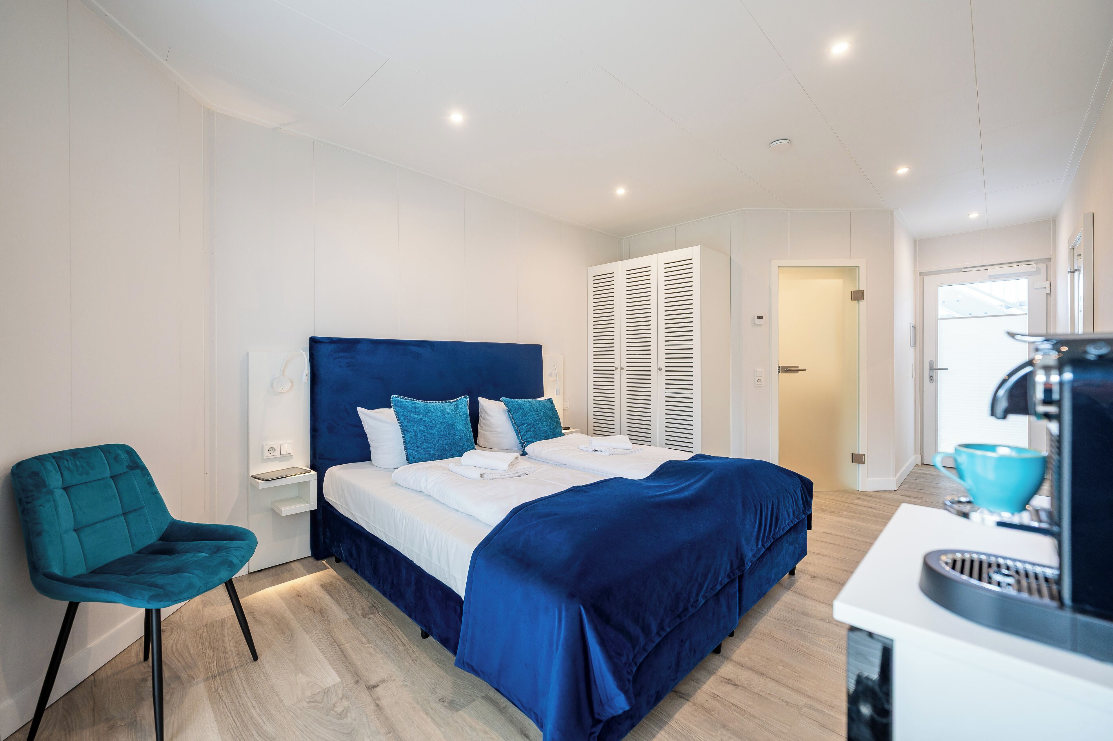 A modern bedroom with a large bed, a blue chair, a bedside table, and a coffee machine.