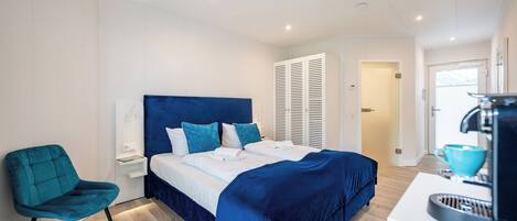 A modern bedroom with a large bed, a blue chair, a bedside table, and a coffee machine.