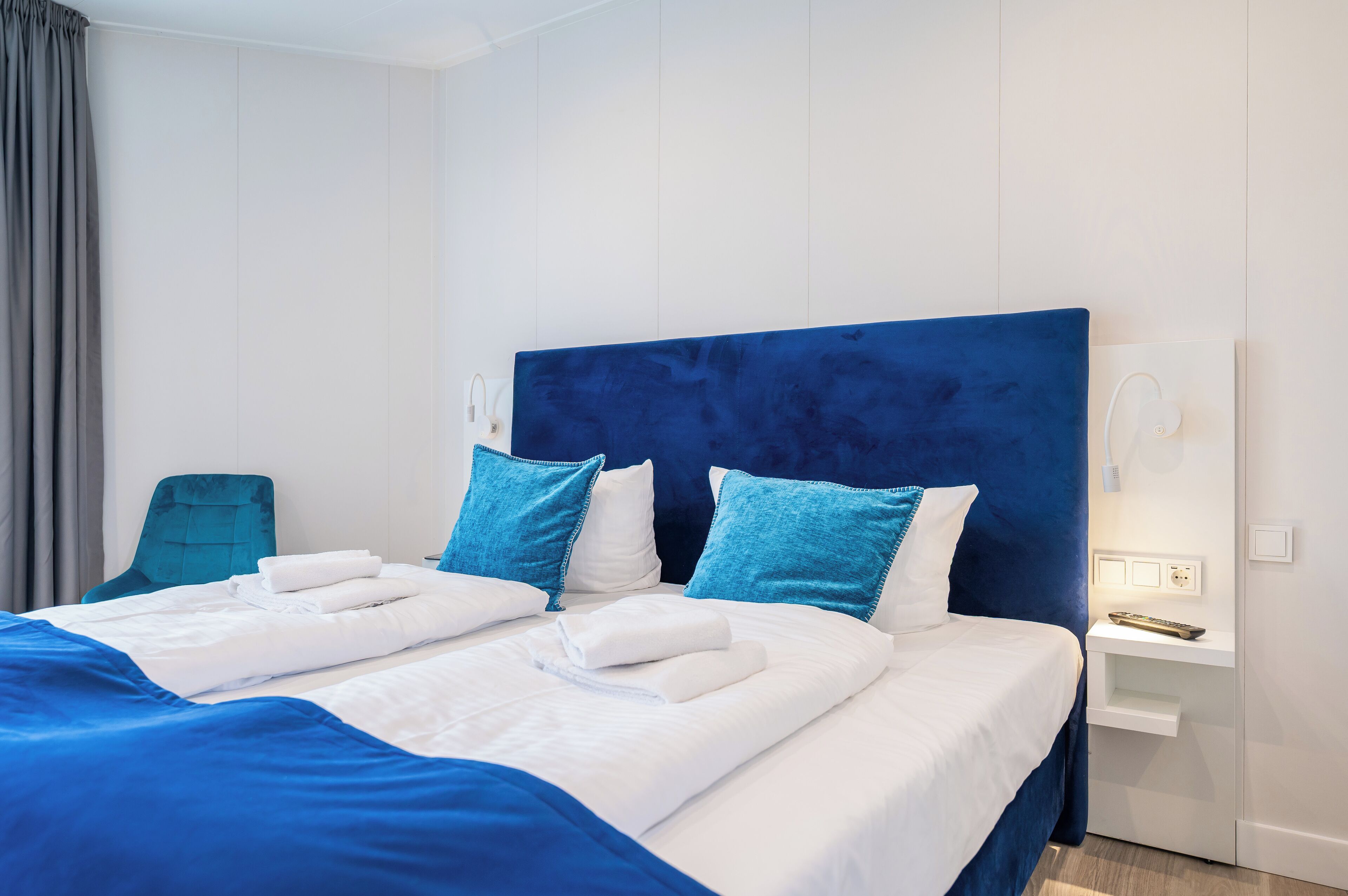 Ein Hotelzimmer mit einem großen Bett, zwei blauen Kissen, einem weißen Kopfteil, einem grauen Sessel und einem Nachttisch mit Fernbedienung.