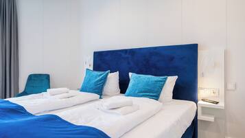 Ein Hotelzimmer mit einem großen Bett, zwei blauen Kissen, einem weißen Kopfteil, einem grauen Sessel und einem Nachttisch mit Fernbedienung.