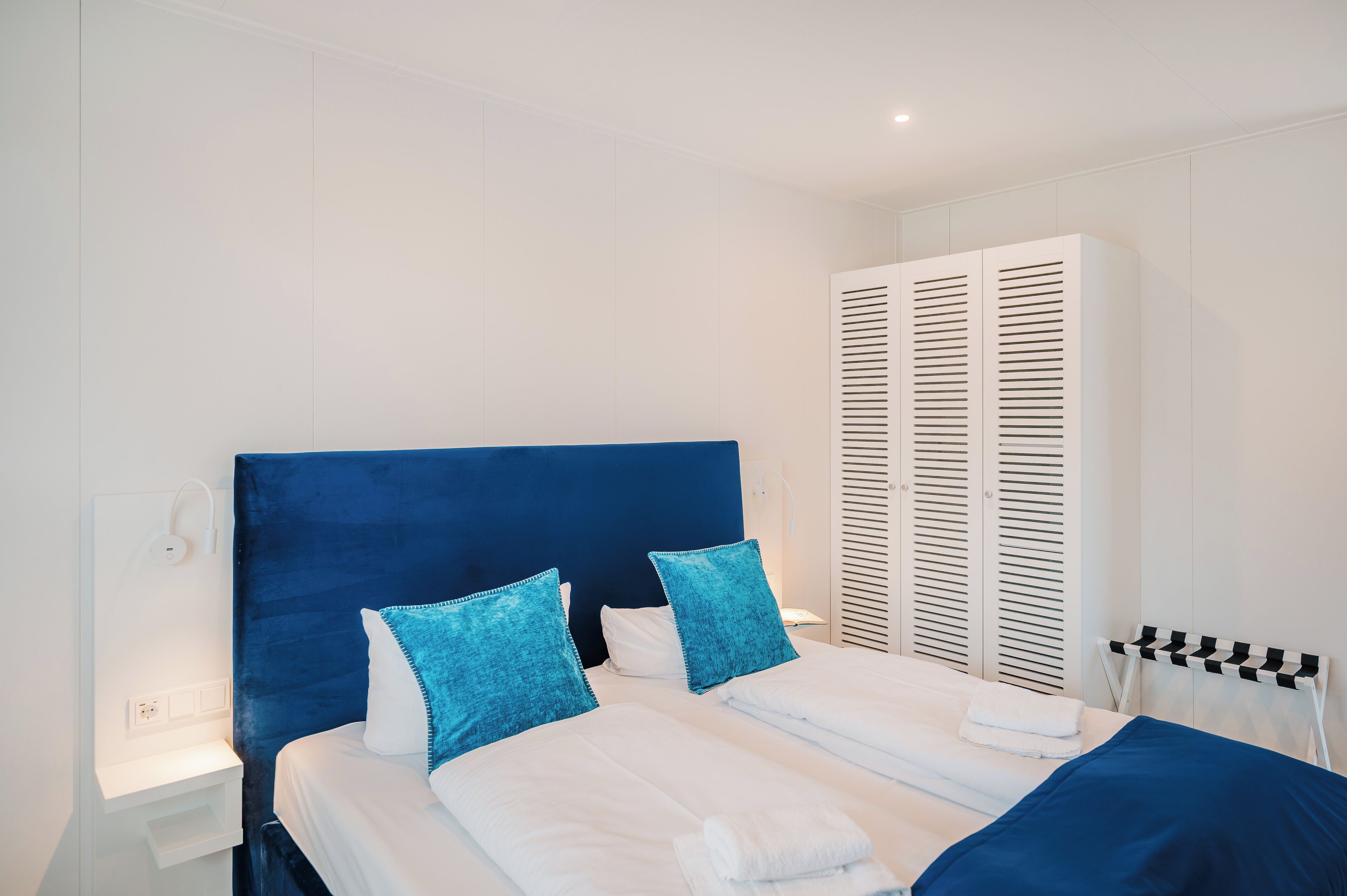 A hotel room with a large bed, white bedding, and a blue headboard. There is a white wardrobe with louvered doors and a white nightstand with a lamp.