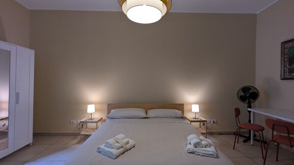 A hotel room with a bed, two bedside lamps, a ceiling fan, and a desk with chairs.