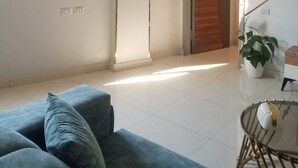 Ein Wohnzimmer mit einem blauen Sofa, einem Glastisch, einem Blumentopf, einer Holztür und einer Wand-Klimaanlage.