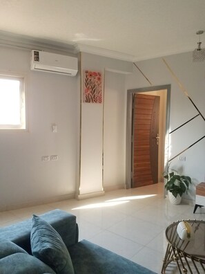 Ein Wohnzimmer mit einem blauen Sofa, einem Glastisch, einem Blumentopf, einer Holztür und einer Wand-Klimaanlage.