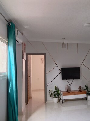 A modern living room with a flat-screen TV mounted on a geometric-patterned wall, a wooden console table with decorative items, and a potted plant.