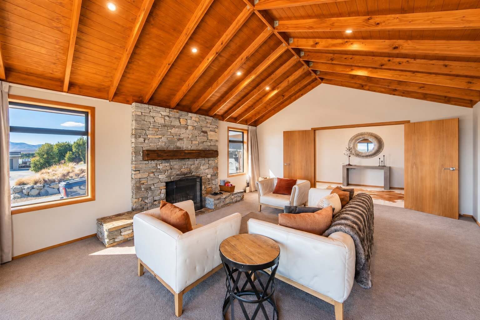 A spacious living area with a stone fireplace, wooden ceiling beams, and a view through large windows.