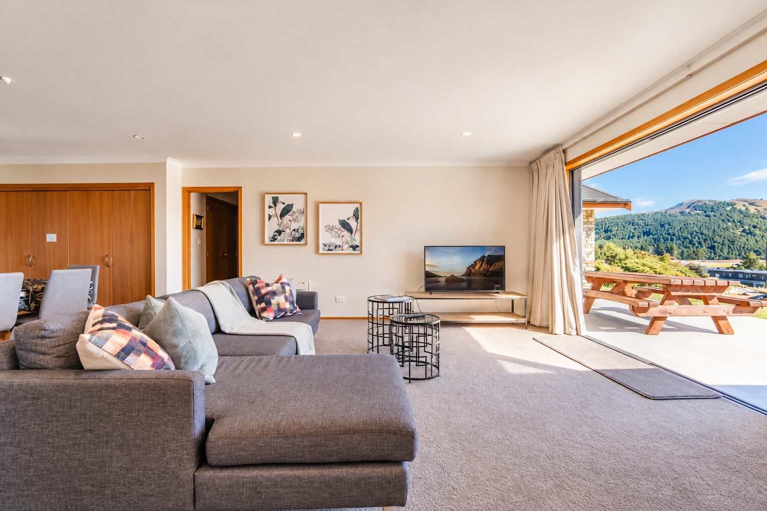 A modern living room with a sofa, a coffee table, and a flat-screen TV. There is a wooden door and a window with a view of a mountainous landscape.