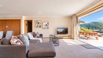 A modern living room with a sofa, a coffee table, and a flat-screen TV. There is a wooden door and a window with a view of a mountainous landscape.