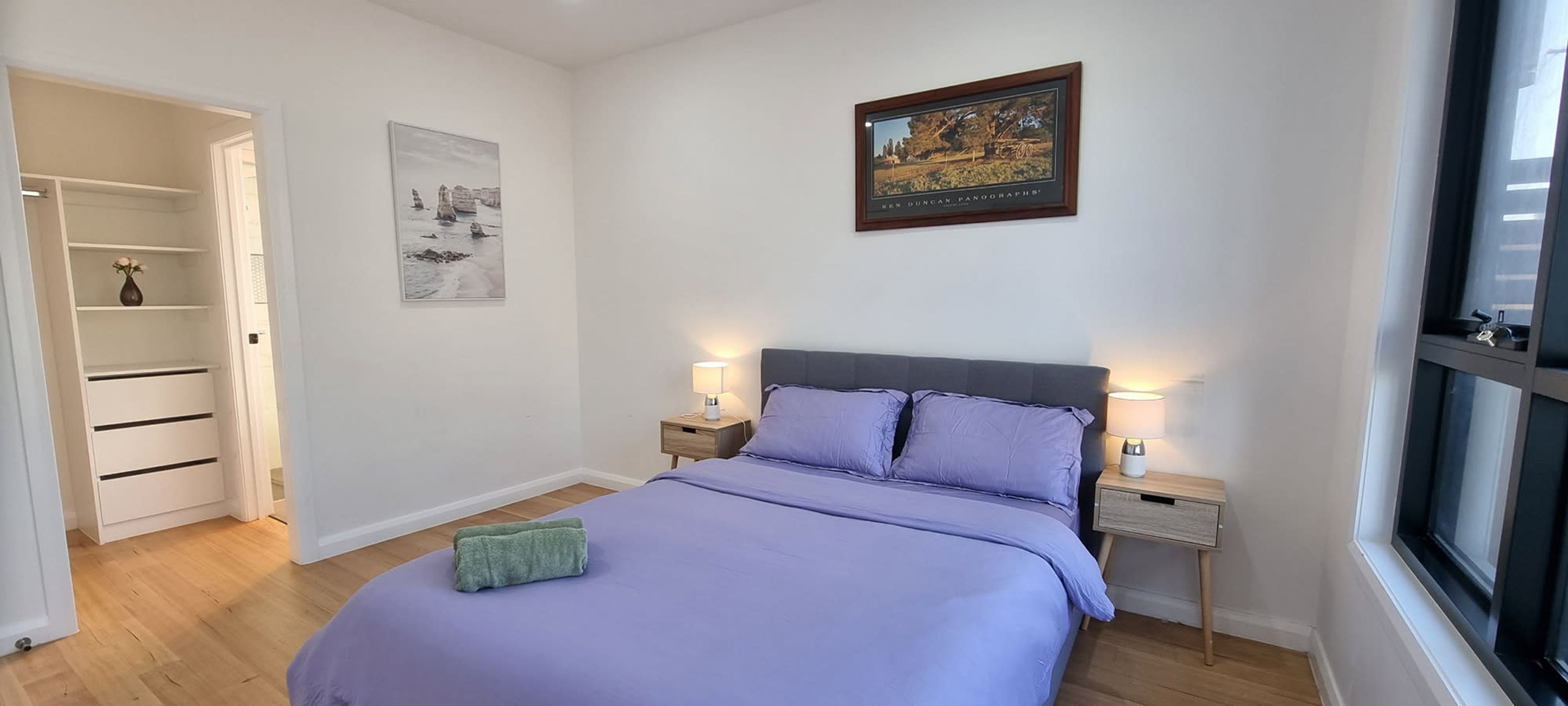 A bedroom with a bed, two bedside tables with lamps, a wardrobe, and a framed picture on the wall.