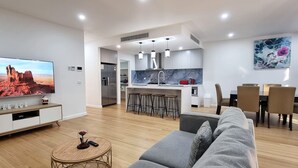 A modern living room with a flat-screen TV, a grey sofa, a small round coffee table, and a dining area with a table and chairs. - Brand new House in Sunshine West (Sunshine West)