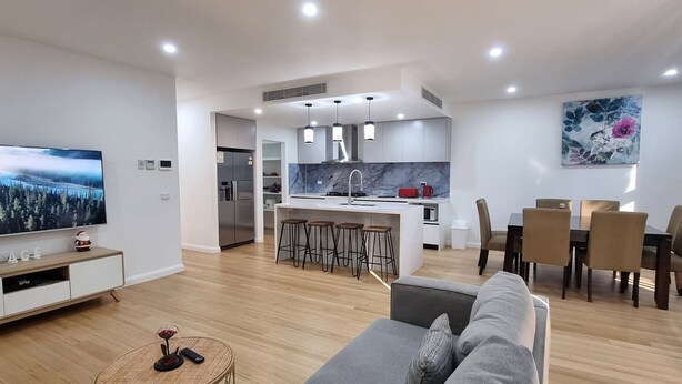 A modern living room with a TV, sofa, and coffee table. A kitchen with a dining area is visible in the background. - Brand new House in Sunshine West (Sunshine West)