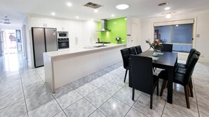 A modern kitchen with white cabinets, a central island, and a dining area with black chairs. - 5 Bedrooms Spacious Home (Sunshine West)