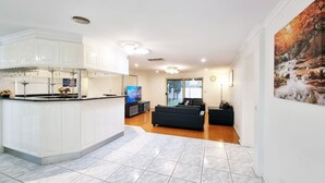 A modern kitchen with white cabinets and a central island, a living area with a black sofa and a TV, and a large window with a view of a tennis court. - 5 Bedrooms Spacious Home (Sunshine West)