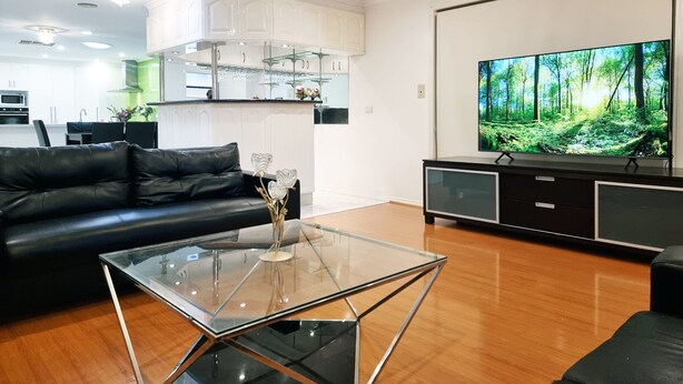 A modern living room with a glass coffee table, a black leather sofa, a flat-screen TV, and a kitchen area in the background. - 5 Bedrooms Spacious Home (Sunshine West)