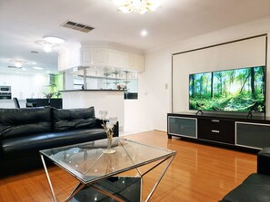 A modern living room with a glass coffee table, a black leather sofa, a flat-screen TV, and a kitchen area in the background. - 5 Bedrooms Spacious Home (Sunshine West)