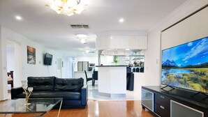 A modern living room with a black leather sofa, a glass coffee table, and a flat-screen TV on a stand. - 5 Bedrooms Spacious Home (Sunshine West)
