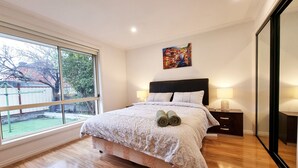 A bedroom with a large window, a bed with rolled towels, a nightstand, and a painting on the wall. - 5 Bedrooms Spacious Home (Sunshine West)