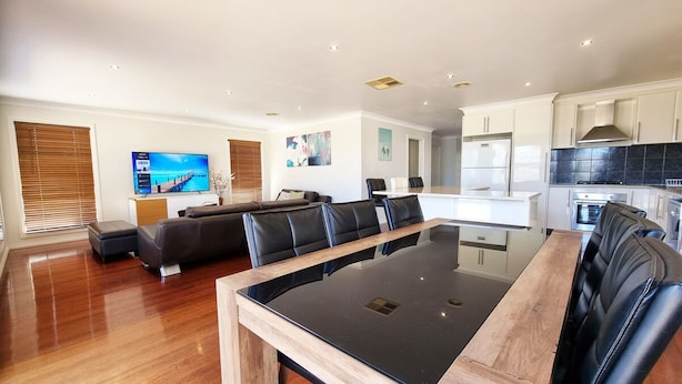 A modern living room with a large TV, a dining area with a glass table, and a kitchen with white cabinets. - St Albans House - Spacious and Modern Home (St. Albans)