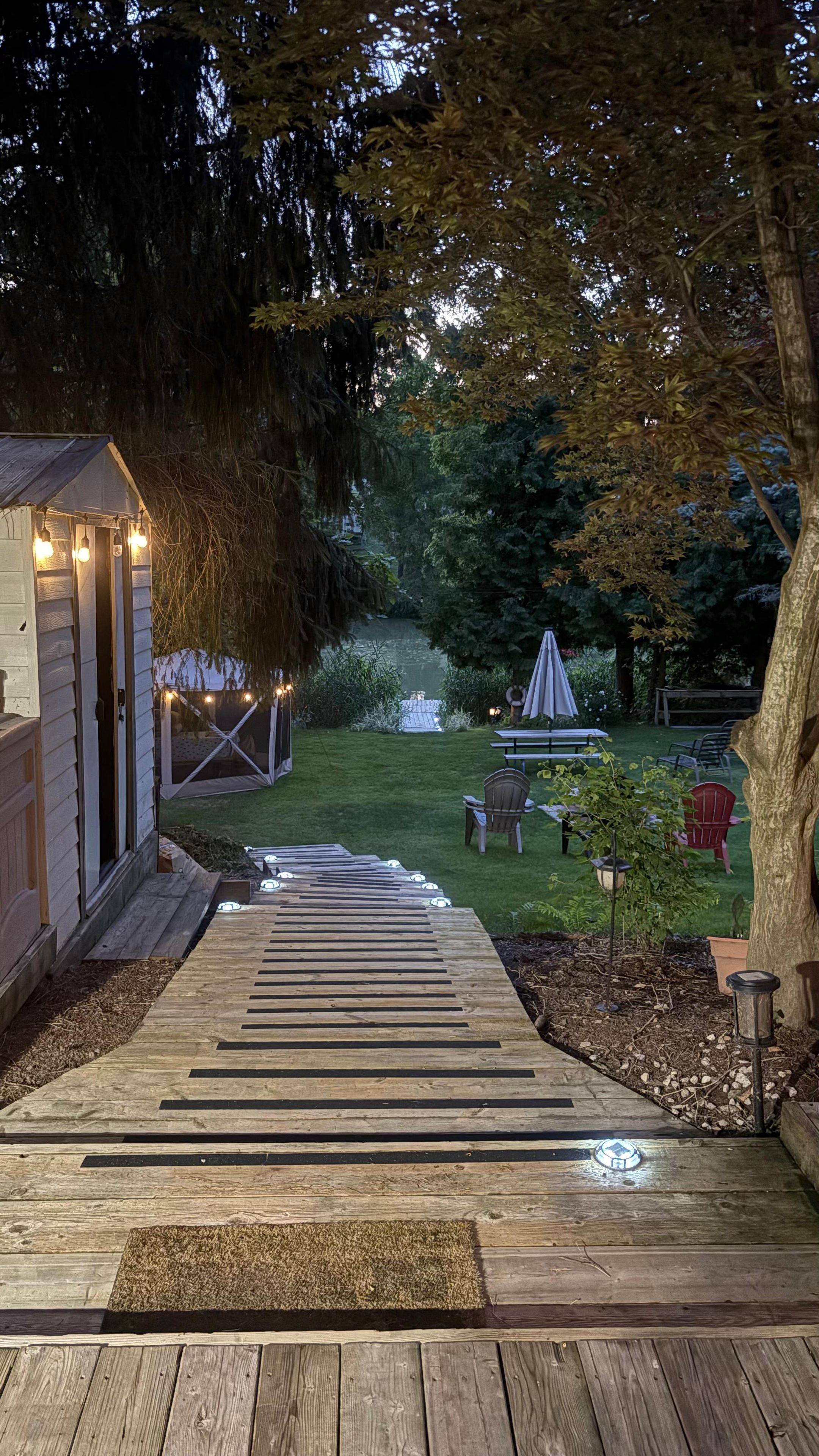 A wooden deck leading to a covered outdoor seating area with string lights, surrounded by trees and a small garden.