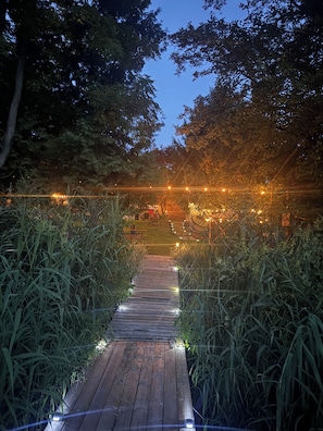 Un sendero de madera bordeado por pastos altos que lleva a un edificio con luces colgantes.