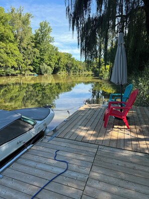 Une jetée en bois s’étendant sur un lac paisible, avec une voiture à l’abri et des chaises colorées sur la jetée.
