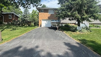A residential driveway leading to a house with a garage, surrounded by trees and a clear sky.