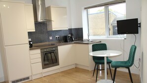 A modern kitchen with white cabinets, a stainless steel range hood, a built-in oven, a coffee maker, and a window with a flat-screen TV mounted on the wall. - East Street Haven - Modern Apartment in Colchester (Colchester)