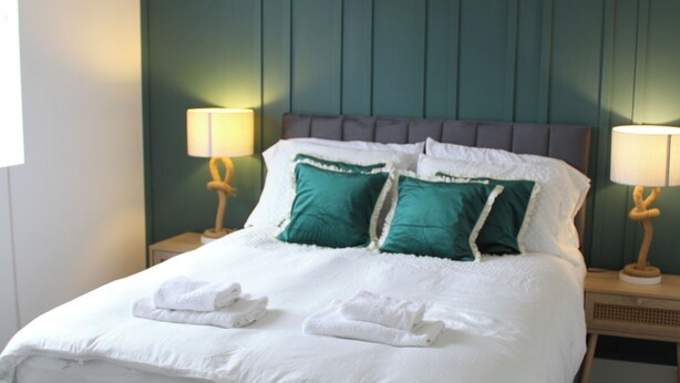 A neatly made bed with white linens and two teal pillows, flanked by two bedside lamps with wooden bases. - East Street Haven - Modern Apartment in Colchester (Colchester)