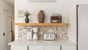Eine Küche mit weißer Arbeitsplatte, einer Kaffeemaschine, einem Toaster, einem Radio und einer Vase auf einem Regal.