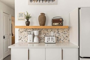 Une cuisine avec un plan de travail blanc, une machine à café, un grille-pain, une radio et un vase posé sur une étagère.