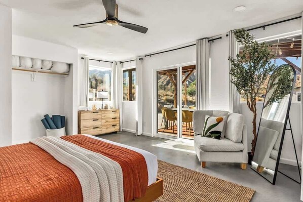 Ein modernes Schlafzimmer mit Bett, Kommode, Stuhl und einem großen Fenster mit Blick ins Freie.