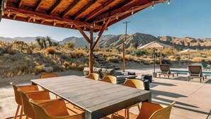 Une terrasse extérieure avec une table en bois et des chaises en osier, entourée d’une végétation désertique et avec des montagnes en arrière-plan.